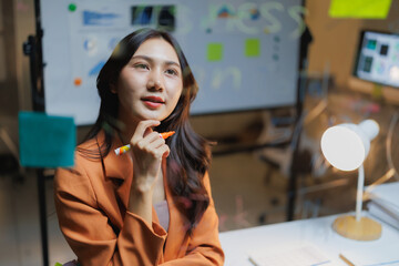 Young Asian businesswoman brainstorming ideas and creative solutions, reflecting on new strategies while working in a contemporary office with data displayed on a monitor