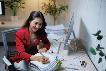 Young asian businesswoman sitting at a desk and writing notes in a pile of documents, actively focusing on paperwork late at night in a modern office environment