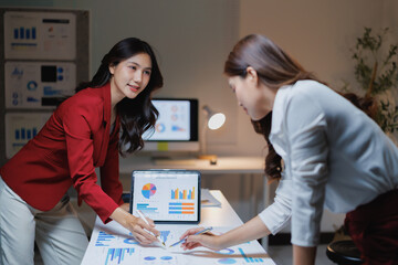 Fototapeta premium Two businesswomen discussing financial reports and pie charts during a late business meeting, analyzing market data and collaborating on a new project using a digital tablet in the office