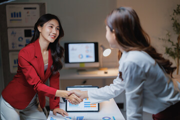 Fototapeta premium Two Asian businesswomen collaborating, sealing a deal with a handshake, showing teamwork and partnership in a modern office environment, with reports and tablet on the desk