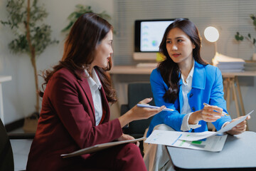 Two professional Asian businesswomen collaborating and analyzing financial charts and reports, working together during a meeting in a modern office environment