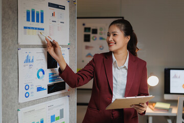 Asian businesswoman discussing financial data and marketing analytics, pointing at a presentation board with various charts and graphs while holding a clipboard in an office setting
