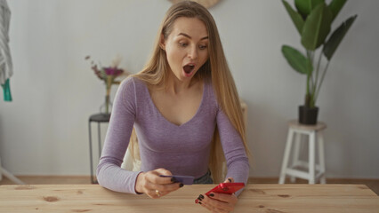 Young woman with long hair excitedly shopping online at home using a smartphone and credit card in...