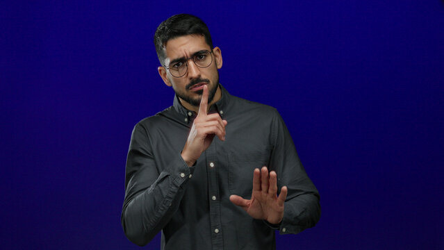 Man wearing glasses gestures quiet with finger over lips and points upward, isolated against blue background, conveying silence and attention.