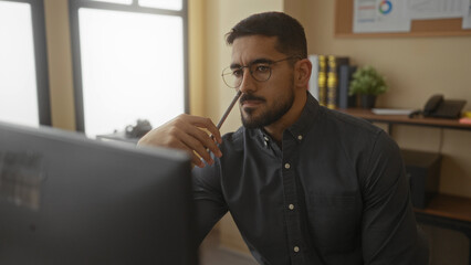 Young man in an office setting contemplates while holding a pencil, surrounded by books and office decor, highlighting a thoughtful and modern workplace environment.