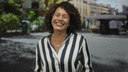 Hispanic middle aged woman in bright striped shirt sticks tongue out on sunlit urban street;...