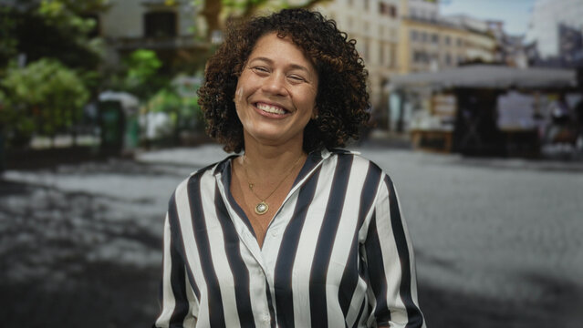 Hispanic middle aged woman in bright striped shirt sticks tongue out on sunlit urban street; playful fun.