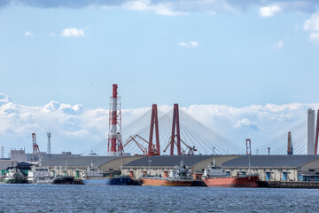 Obraz premium Industrial port scene featuring cargo ships docked along quay with warehouses, and cranes and red cable-stayed bridge visible in background.