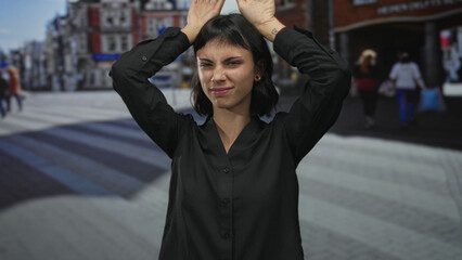 Fototapeta premium Young hispanic woman makes ear gesture with hands on head in urban street scene wearing black shirt; playfulness.
