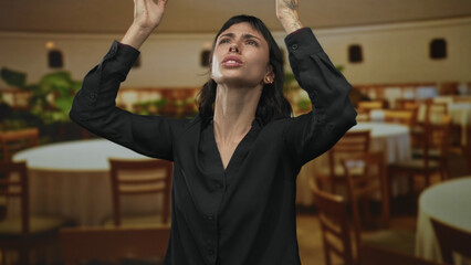 Young hispanic woman wearing black blouse covers ears with hands among tables in restaurant; frustration.