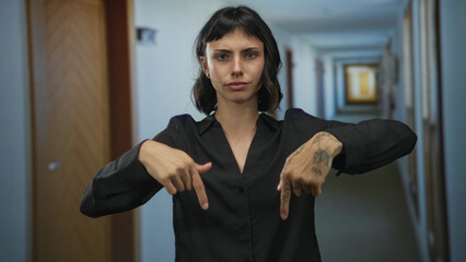 Young hispanic woman pointing finger downward in hotel building corridor with wooden doors behind her; alertness warning.