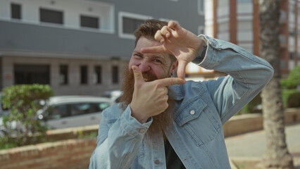 Man with long beard and denim jacket framing face with fingers on sunlit city street; creative vision.