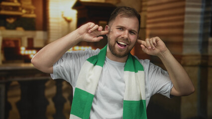 Obraz premium Man wearing green and white scarf plugs ears with fingers and smiles inside a church building, caucasian young fan in white tshirt; defiance.