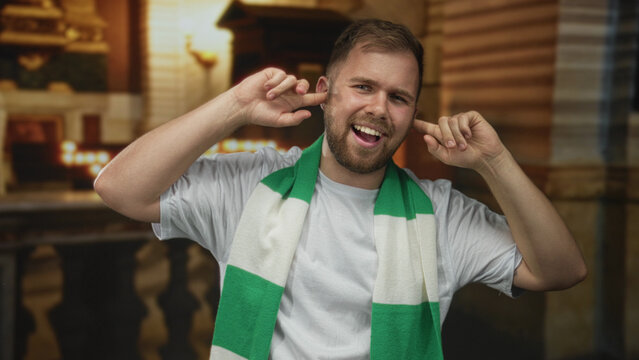 Man wearing green and white scarf plugs ears with fingers and smiles inside a church building, caucasian young fan in white tshirt; defiance.