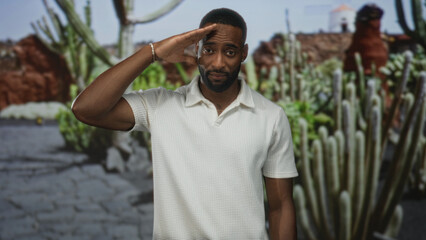 Man saluting with hand to forehead in park among cactus plants and paved path, white polo visible;...