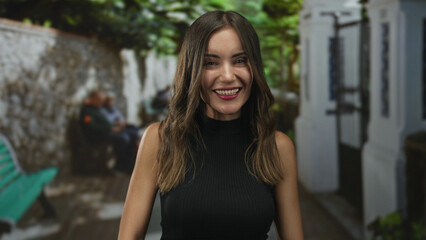 Woman giving thumbs up and smiling in a street courtyard with green foliage, white building and bench, wearing a black sleeveless top; joy approval.