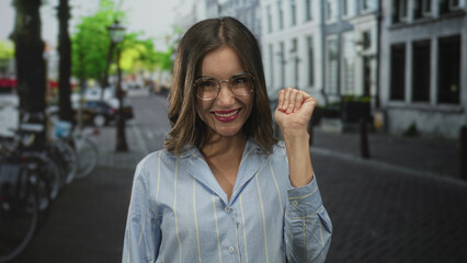 Fototapeta premium Woman with glasses smiling and puckering lips wearing blue striped shirt on a city cobbled street near parked bicycles and white buildings; playful confidence.