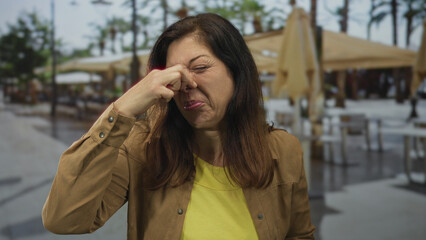 Fototapeta premium Woman pinches nose on street terrace outside a restaurant with palm trees and umbrellas while grimacing; disgust.