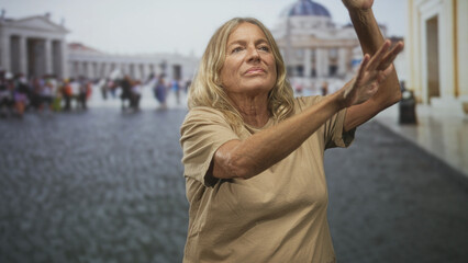 Senior blonde woman raising both arms with bare forearms visible in a busy piazza before a basilica, looking upward  quiet contemplation. © Krakenimages.com