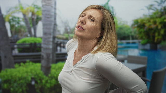 Middle age woman holds lower back and leans backward in green park by poolside chairs on sunny morning; discomfort.