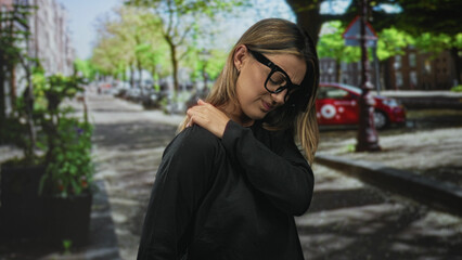 Fototapeta premium Blonde woman rubbing her shoulder on a leafy street wearing dark top and glasses near a parked car and planter for backache; discomfort.