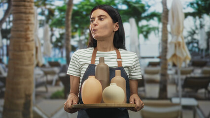 Woman potter holds tray of brown ceramic pots with hands while wearing apron in studio workshop by...