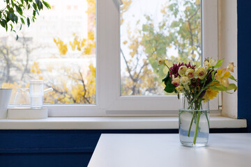 A bouquet of flowers sits on a windowsill near a glass jar overlooking a sunny day © Tetiana Romaniuk
