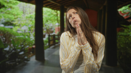 Woman with hands pressed together in pleading pose in a hotel corridor with pillars and greenery; anxious hope.