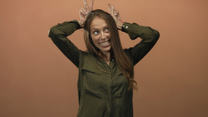 Woman making bunny ears with fingers and smiling in studio with peach backdrop wearing olive shirt; playful joy fun.
