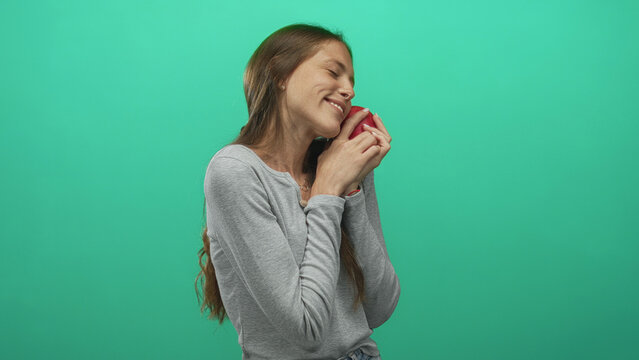 Woman holding red apple to cheek in studio with teal green background, eyes closed and gentle smile while embracing the fruit; contentment affection.