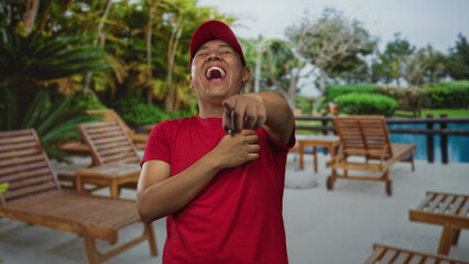 Fototapeta premium Young man in red cap and red shirt pointing and laughing with hand on chest at building near poolside lounge chairs; playful joy.