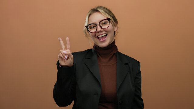 Woman smiling while making a victory sign with fingers, standing against a brown wall, wearing glasses and a blazer, showcasing a confident and cheerful expression.