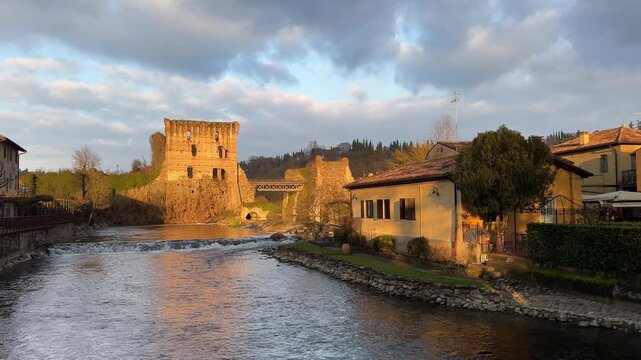 Golden hour view of Borghetto sul Mincio's Ponte Visconteo bridge and medieval ruins reflecting in calm river waters, highlighting historic Italian architecture and serene landscape.