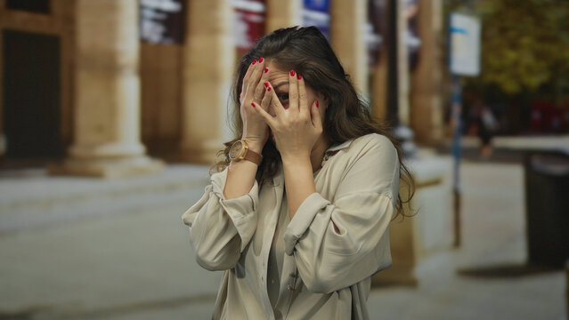 Young hispanic woman pressing palms to cheeks with furrowed brow and open mouth in an old town street; fear.