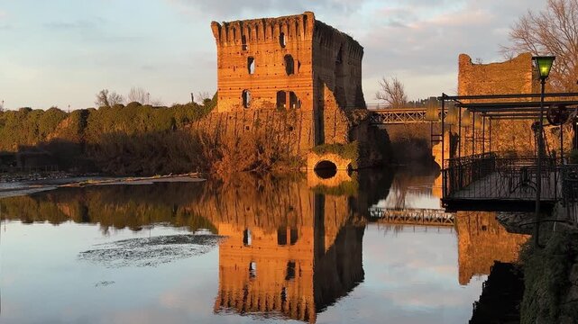 Golden hour view of Borghetto sul Mincio's medieval ruins and Ponte Visconteo bridge reflecting in the calm river water, showcasing historic Italian architecture and scenic riverside dining.
