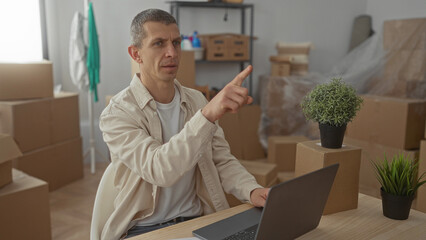 Fototapeta premium Man in casual attire working on laptop surrounded by cardboard boxes and plants in new apartment living room setting with natural light streaming in through windows.
