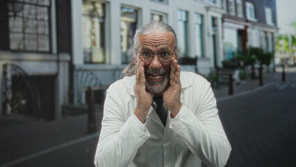 Man scientist bearded with gray long hair in white lab coat with hands cupping cheeks on street in city; curiosity surprise.