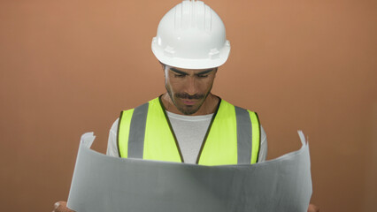 Man holds rolled blueprint with both hands while wearing safety vest and white hardhat in brown studio; strategic planning confidence.