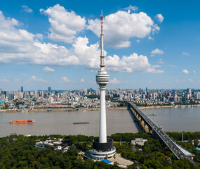 Obraz premium Wuhan TV Tower stands majestically by the Yangtze, surrounded by a bustling cityscape and serene river traffic, under a summer sky dotted with clouds. The tower, a beacon of technology, with sleek, fu