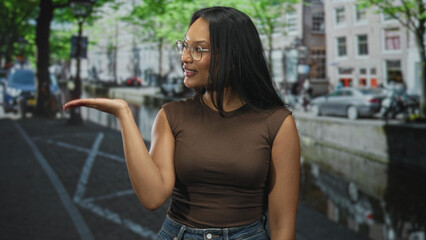 Fototapeta premium Woman points finger up on a canal street with glasses and brown top, presenting gesture by parked cars and canal water; confidence invitation.