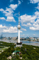 Obraz premium Wuhan TV Tower stands majestically by the Yangtze, surrounded by a bustling cityscape and serene river traffic, under a summer sky dotted with clouds. The tower, a beacon of technology, with sleek, fu