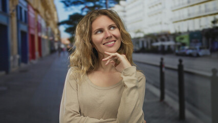 Fototapeta premium Woman hand touching lips while she poses on a street with blonde hair and a smiling expression in a playful casual portrait on a sidewalk.
