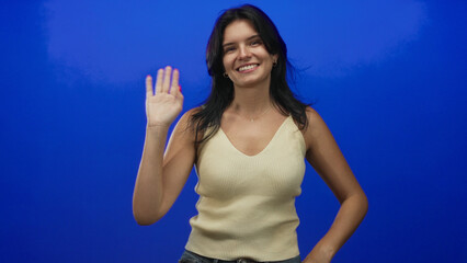 Woman waving hand with smiling expression wearing beige tanktop and blue jeans in blue studio; friendly greeting.