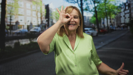 Fototapeta premium Older woman making ok sign with hand over eye on a city street by a canal, smiling and wearing a green blouse near parked cars; playful joy.