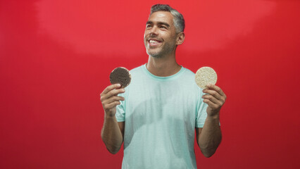 Man holding two corn pastries in hands, smiling and presenting a chocolate biscuit and a rice cake against a red studio wall; choice contentment.