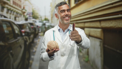 Man doctor in white coat holds a model brain in his palm on an urban street beside a building;...