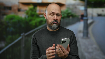 Man outdoors with smartphone on urban street showcasing thoughtful expression while dressed casually against blurred city backdrop captured in natural light setting.