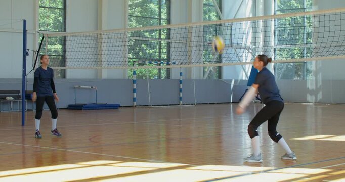 Girls pass the ball to each other, practicing overhead and underhand serves. Two female volleyball players come to the court with a ball and start warming up.