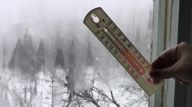 Outdoor thermometer in a woman's hand with raindrops in the background of a raindrop window. High humidity outside and in the apartment