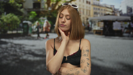 Fototapeta premium Blonde woman with tattoos and sunglasses standing thoughtfully on a busy urban street with outdoor market.